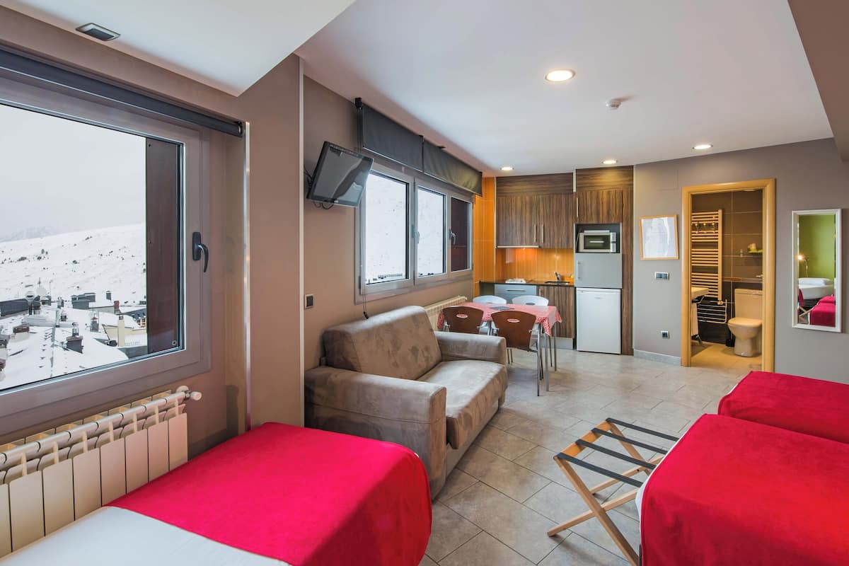 A modern hotel room with a large window, a sofa, a dining table, and a kitchen area.