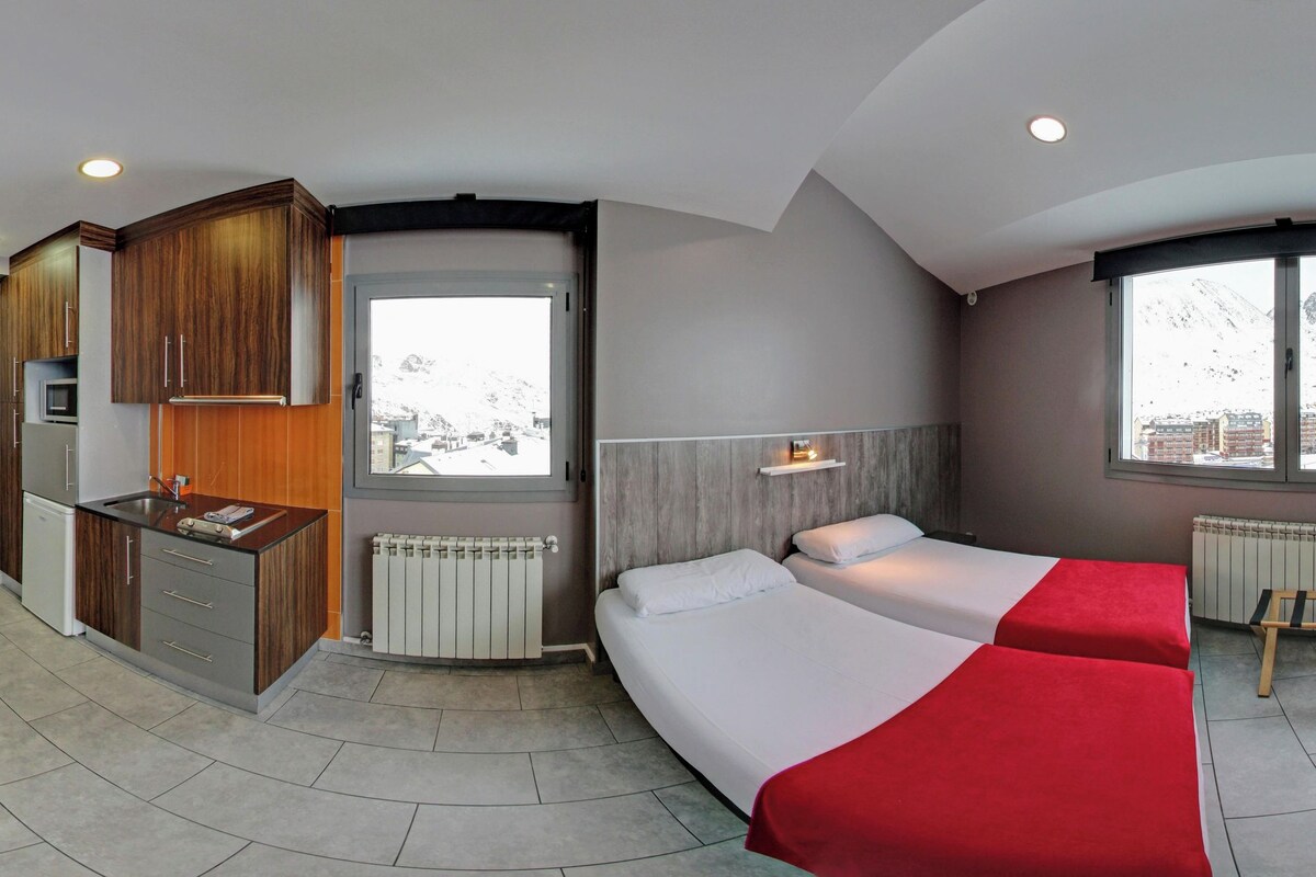 Quarto de hotel com cozinha, duas camas, uma mesinha e uma televisão.