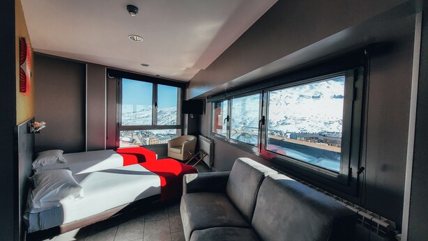 Una habitación de hotel moderna con cama, sofá, televisión y ventanas amplias que dan a una vista nevada de la ciudad.