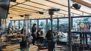 Un área de estar al aire libre con mesas de madera y sillas de metal, ventanas amplias y vistas panorámicas de la ciudad.