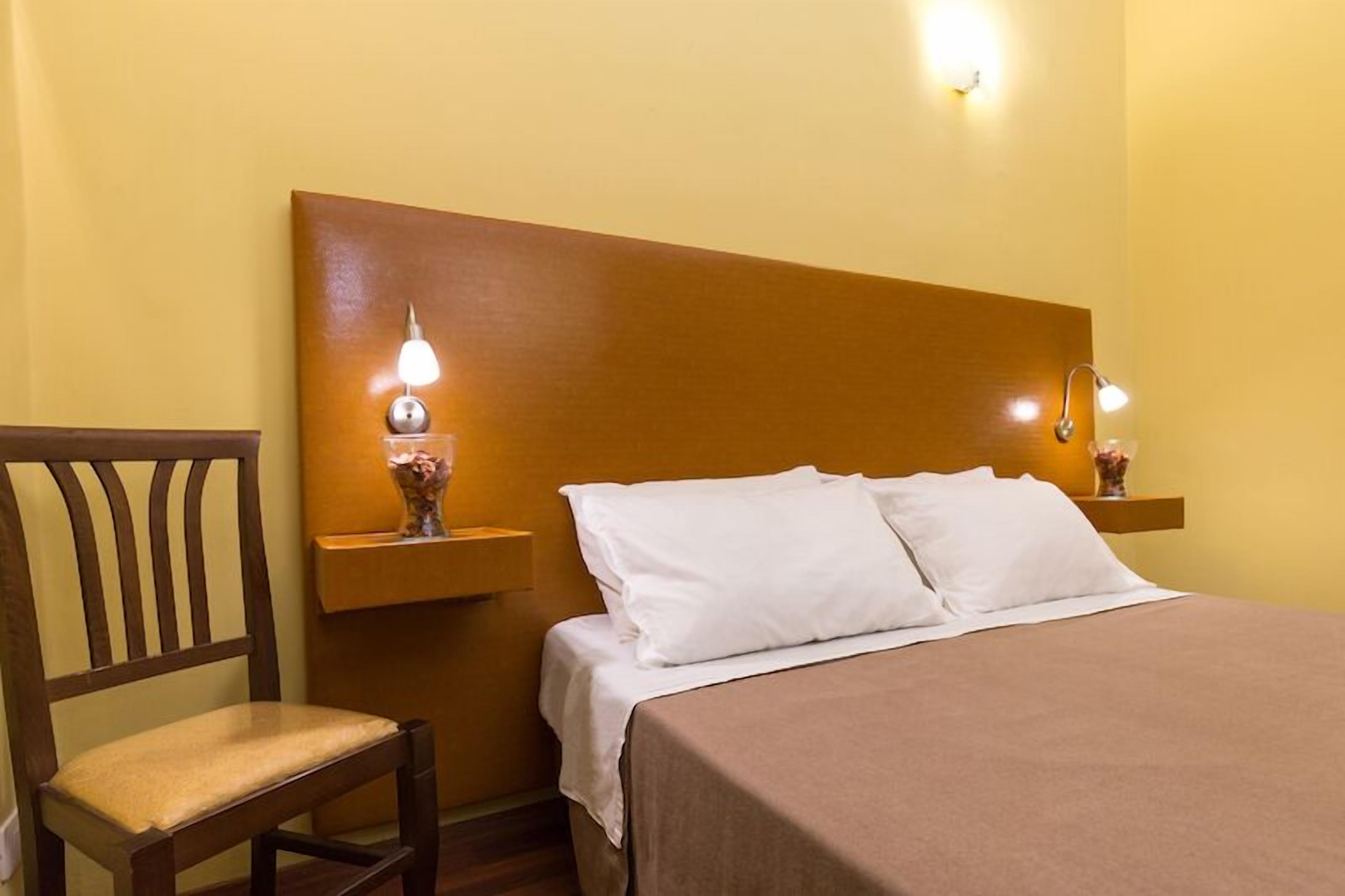 Ein Hotelzimmer mit einem Holzkopfteil, einem Einzelbett mit weißen Leinen, zwei Nachttischlampen und einem kleinen Tisch mit einer Vase.