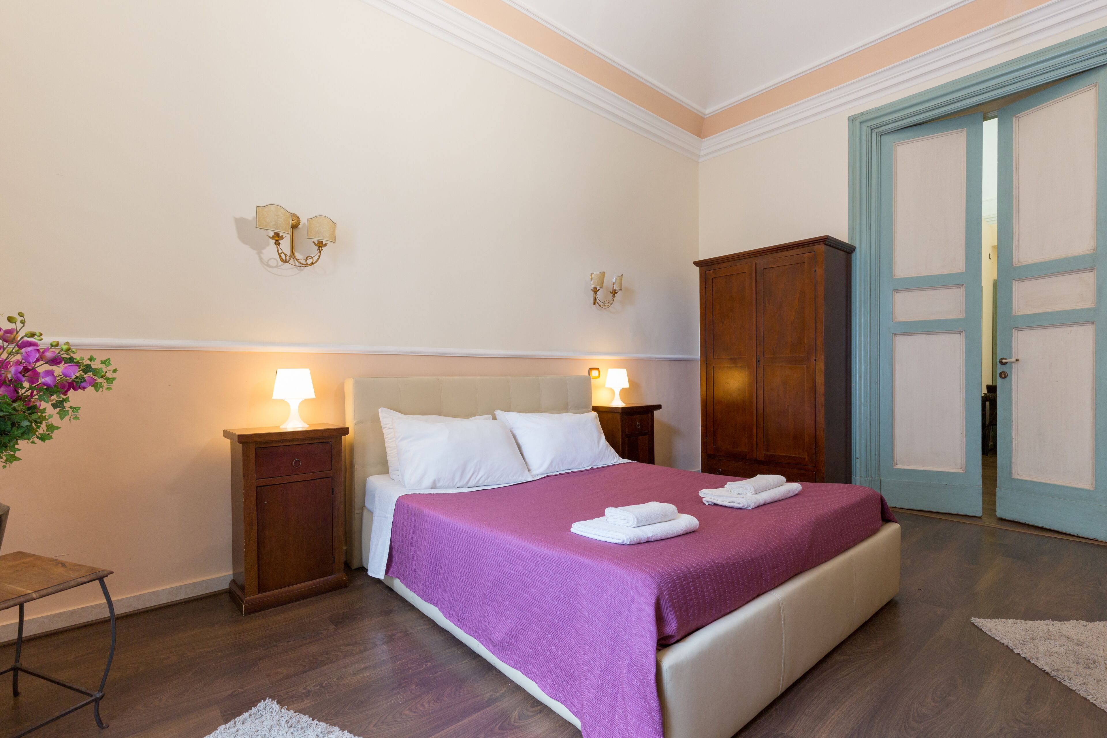 Une chambre d’hôtel bien rangée, avec un lit, des tables de chevet, une petite table, une armoire en bois et une porte.