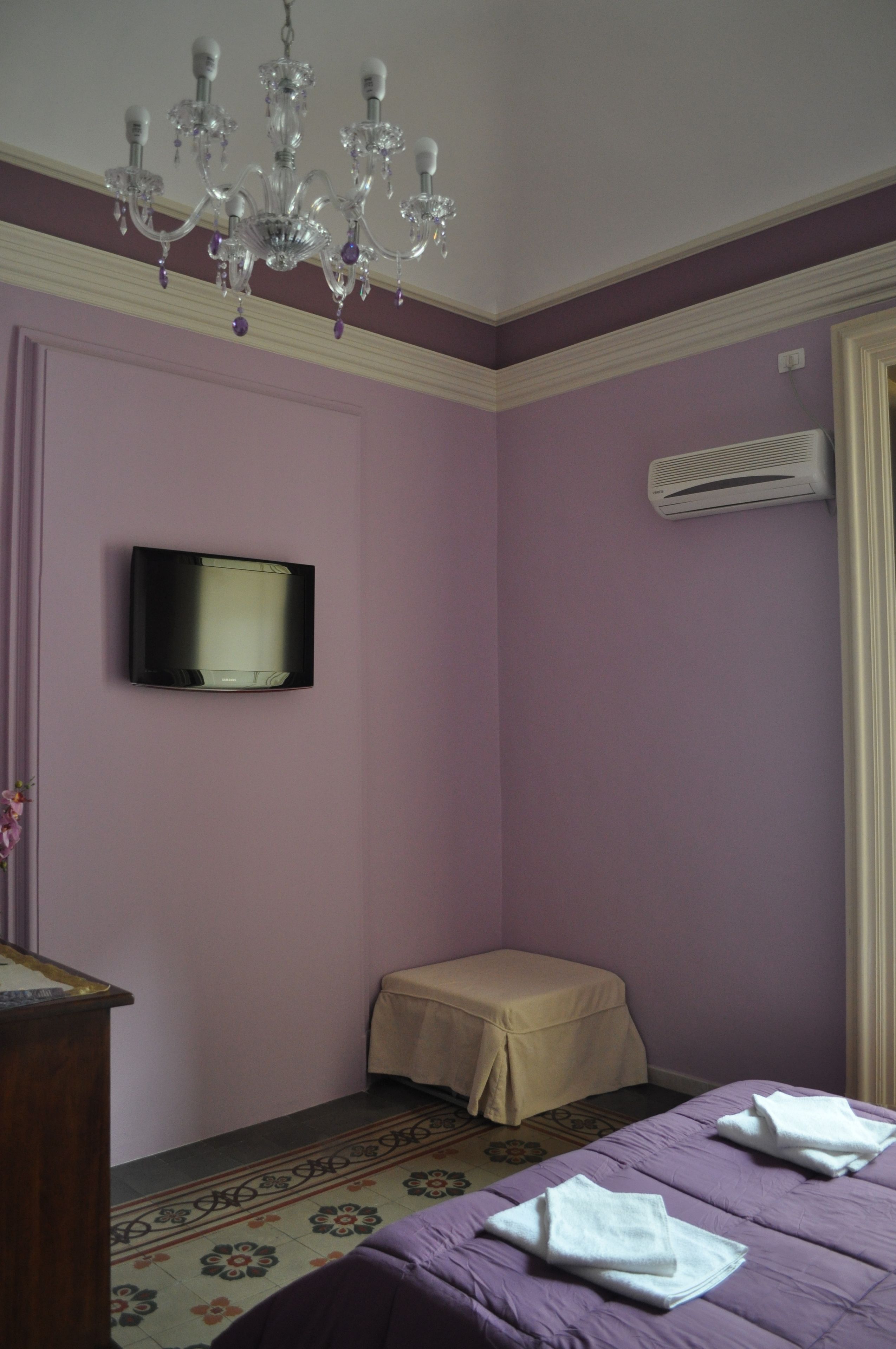 A room with a bed, a television mounted on the wall, a chandelier, and an air conditioning unit.