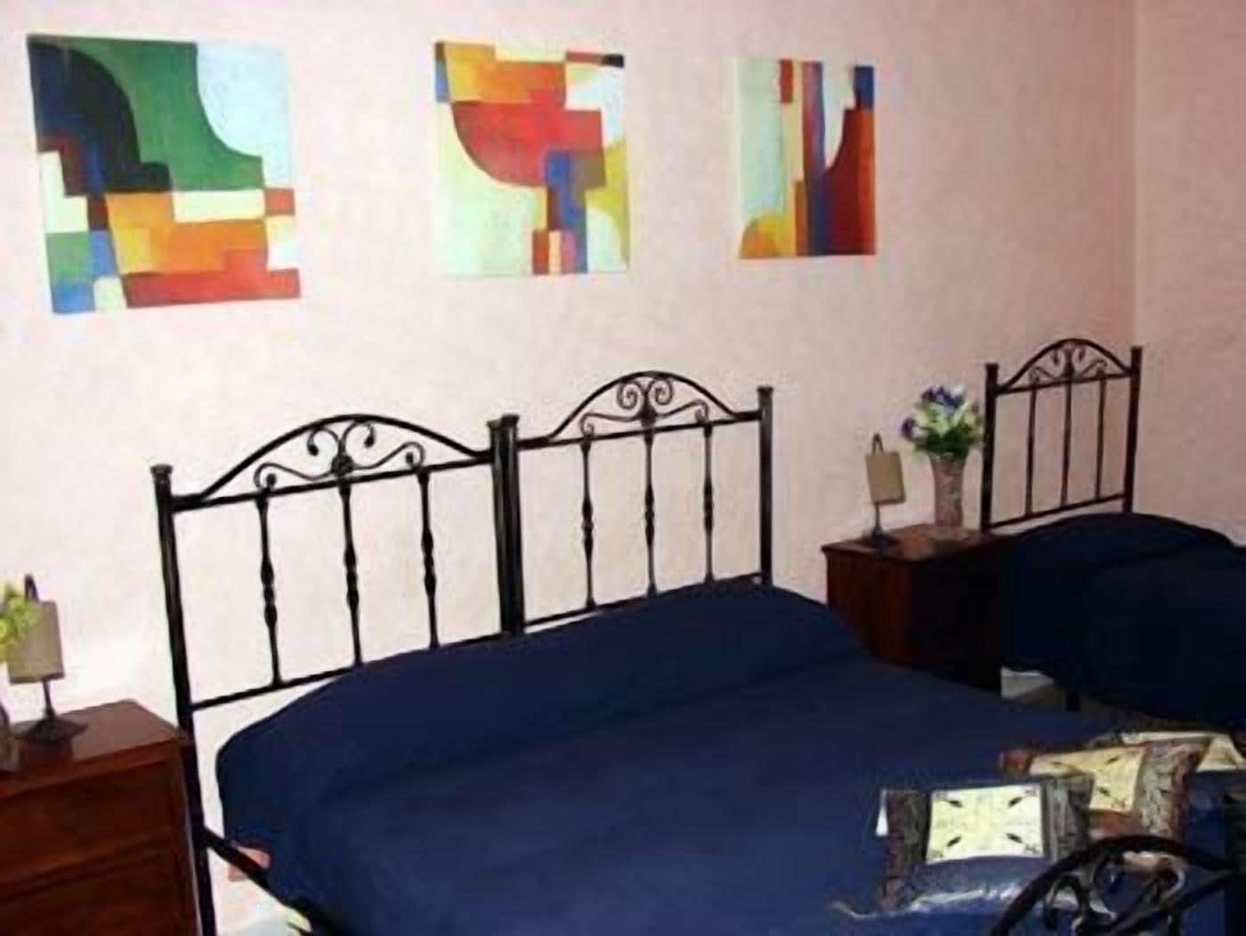 A bedroom with a metal bed frame, a blue bedspread, and three abstract paintings on the wall.
