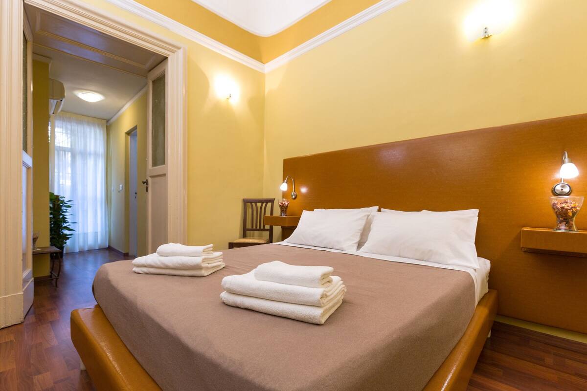 Habitació d'hotel amb un llit gran, un panell de capçalera de fusta i una tauleta de nit amb una làmpada.