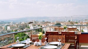 Uma área de refeições com mesas e cadeiras, vista para um horizonte urbano e um corrimão.
