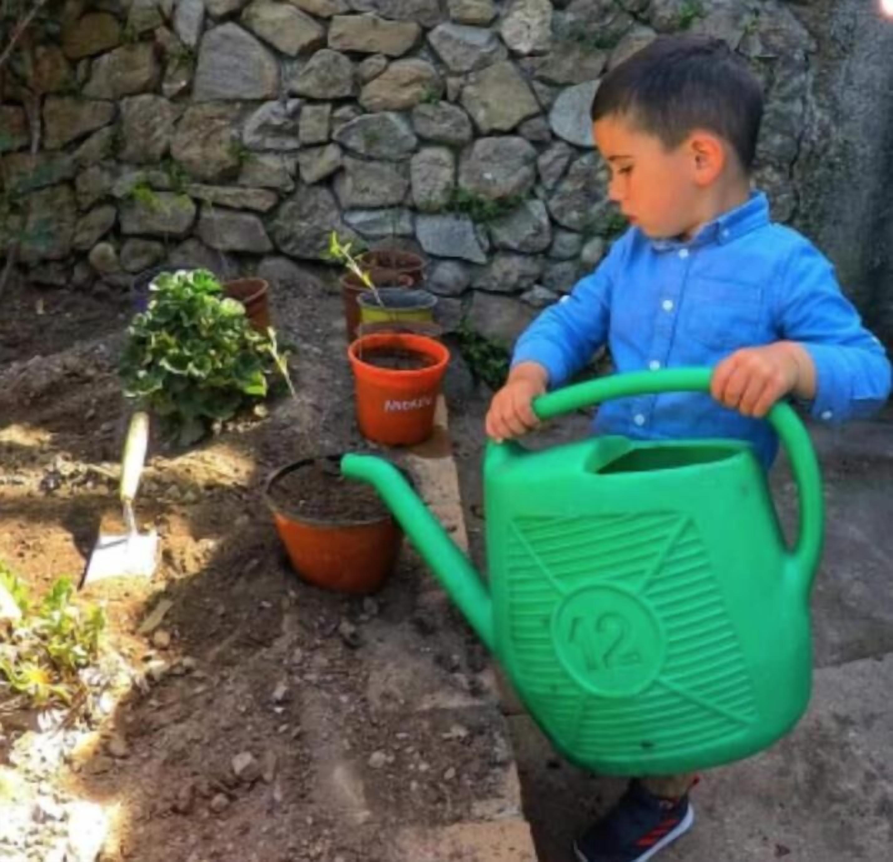 A child holding a green watering can in a garden with potted plants and a stone wall.