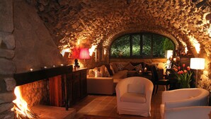 Ein gemütlicher Raum mit Steinwänden, einem Kamin, bequemen Sitzmöglichkeiten und Blick durch ein großes, rundbogenförmiges Fenster.