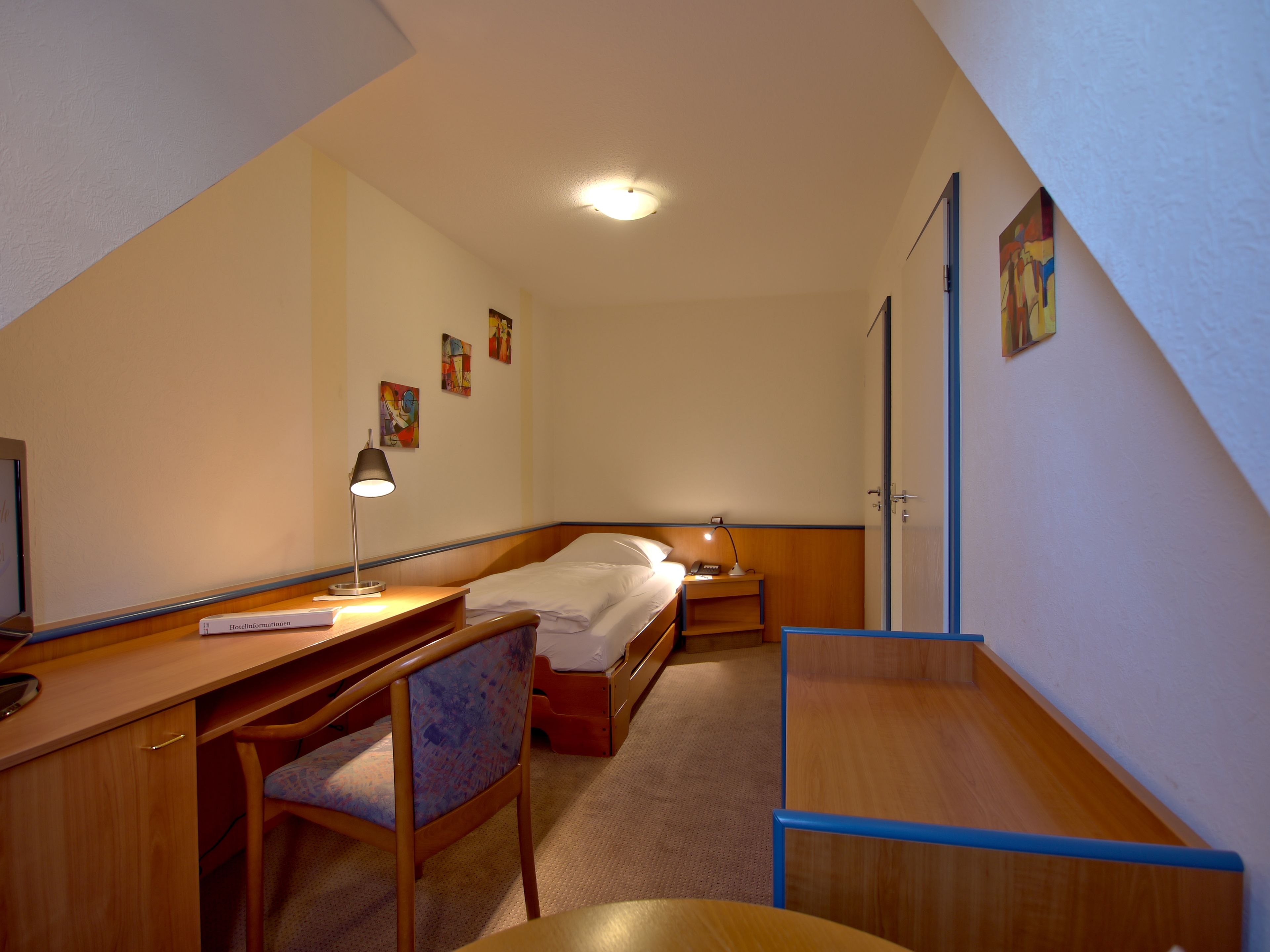 A hotel room with a bed, desk, chair, and a small table. There are two bedside tables with lamps, a wall-mounted TV, and a door with a handle.