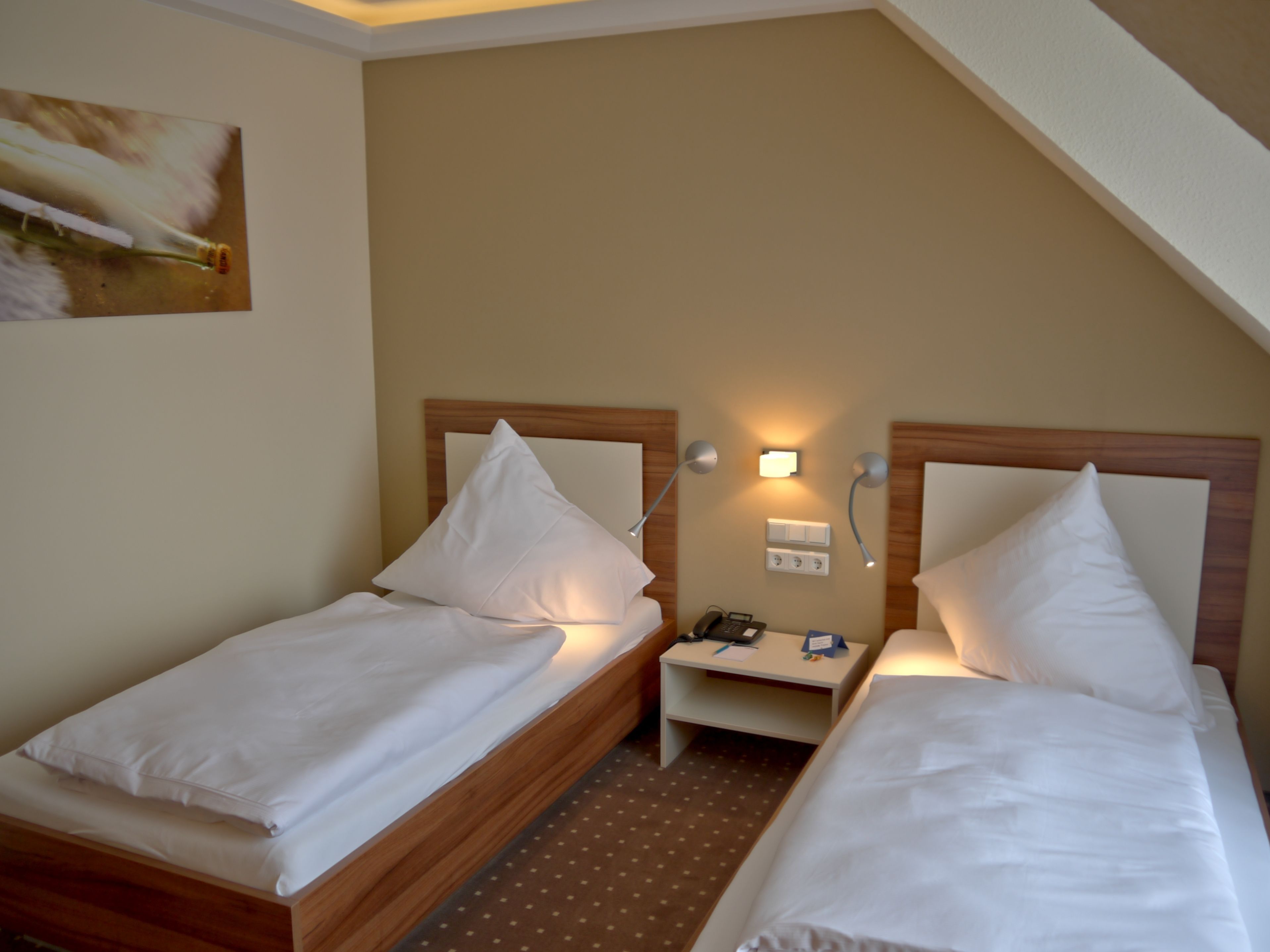 A small hotel room with two single beds, a nightstand with a lamp, a phone, and a wall socket.