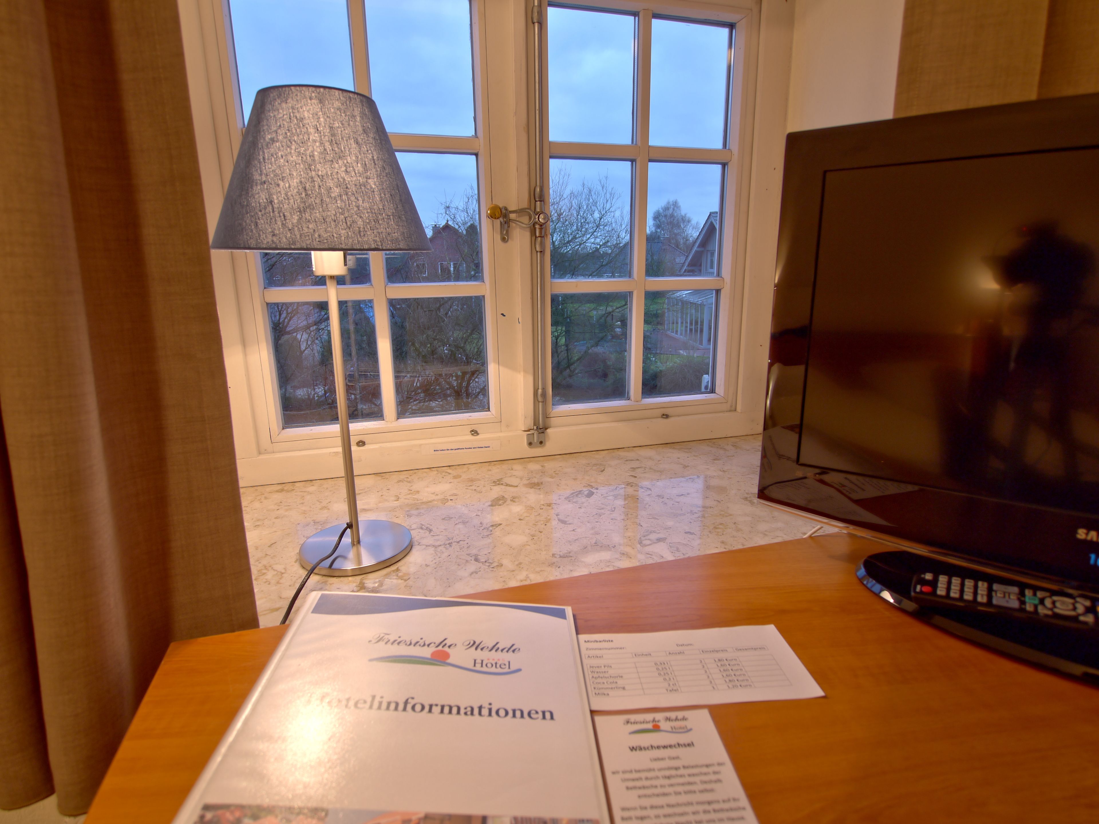 A hotel room with a large window, a lamp, a television, and a table with hotel information and a menu.