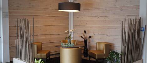 Un área de recepción con pared de madera, un mostrador central con un letrero que dice 'WILLKOMMEN', dos sillas y una lámpara colgante.