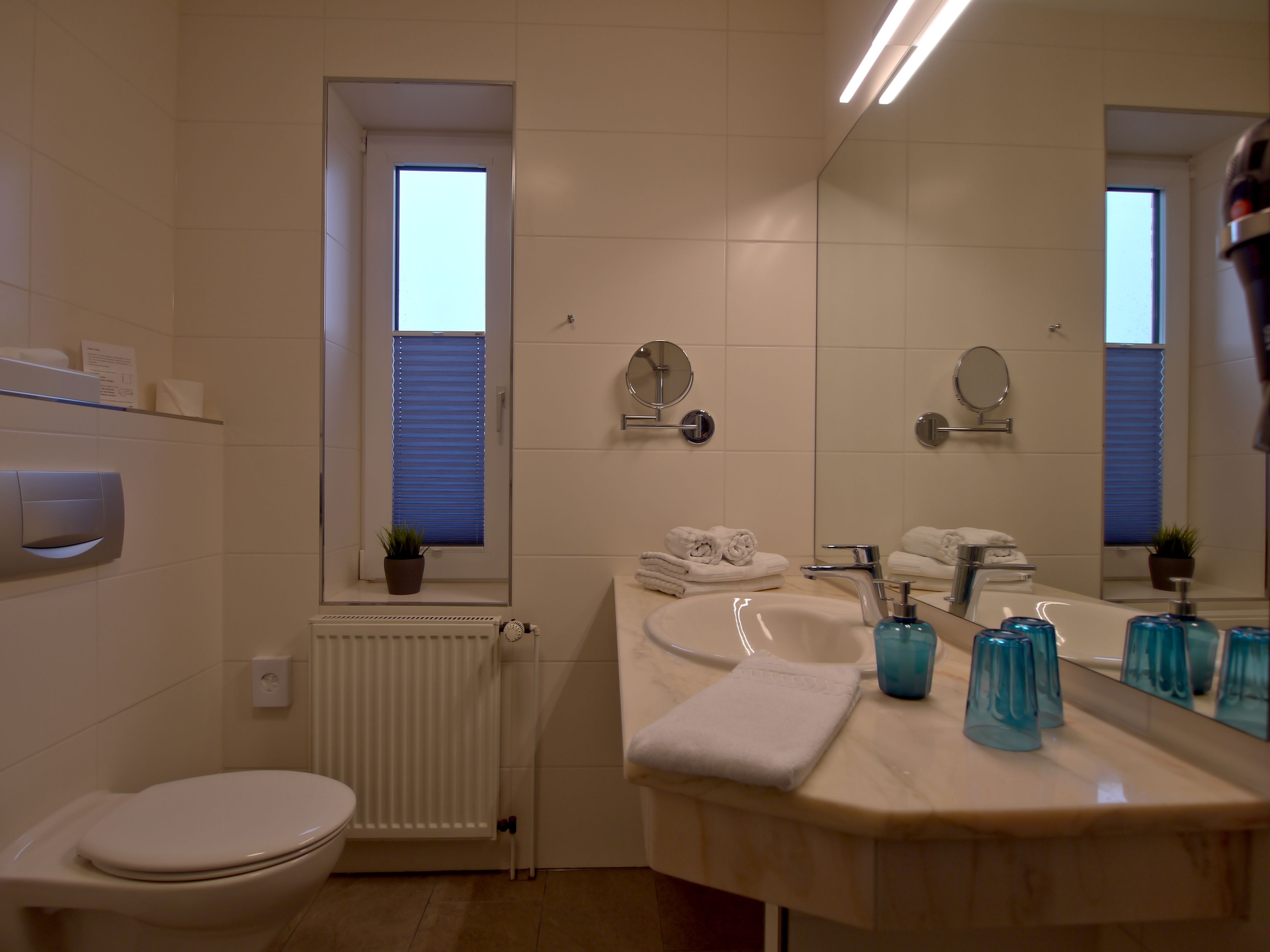 Un baño moderno con inodoro, lavamanos y un espejo grande. Hay toallas y artículos de aseo en la encimera.