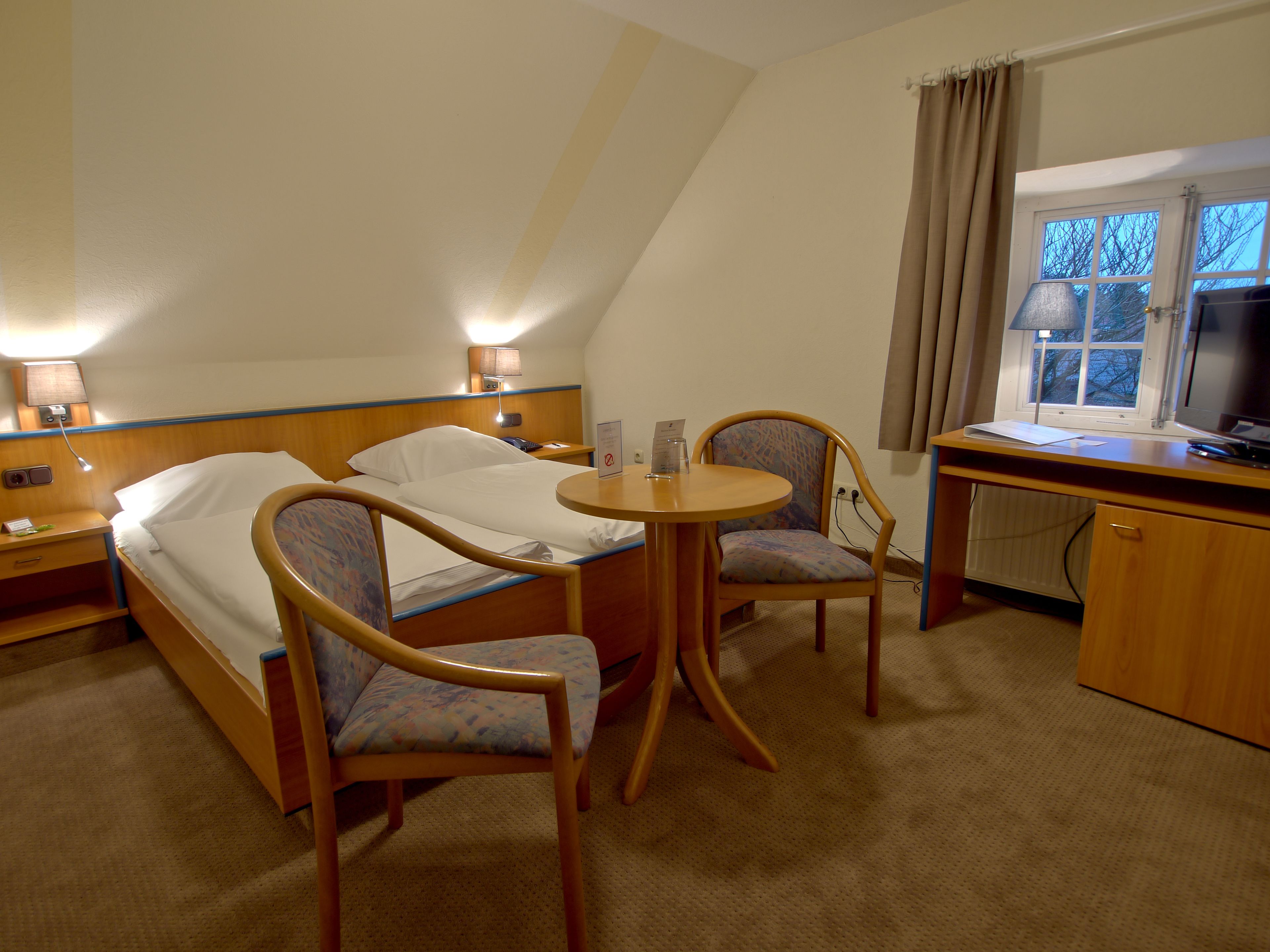 A hotel room with a bed, two chairs, a desk, a lamp, and a television.