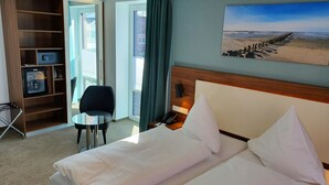 A hotel room with a bed, a desk, a chair, a minibar, and a painting of a beach. - Hotel Friesische Wehde (Bockhorn)