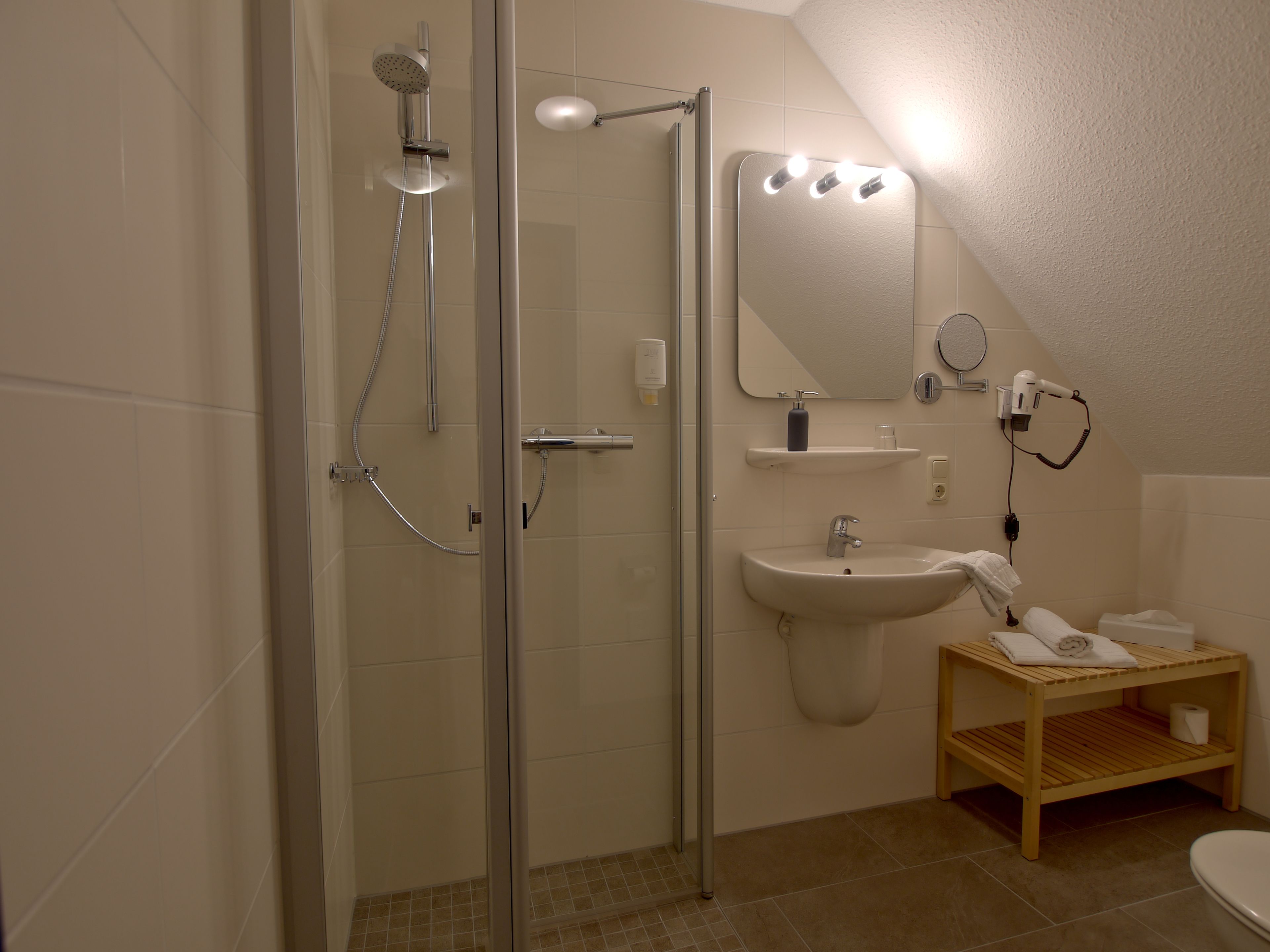 Un baño con ducha envuelta en vidrio, lavamanos con espejo y estante de madera con toallas.