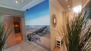 A hallway with a beach mural, a small white stool, and tall plants. - Hotel Friesische Wehde (Bockhorn)