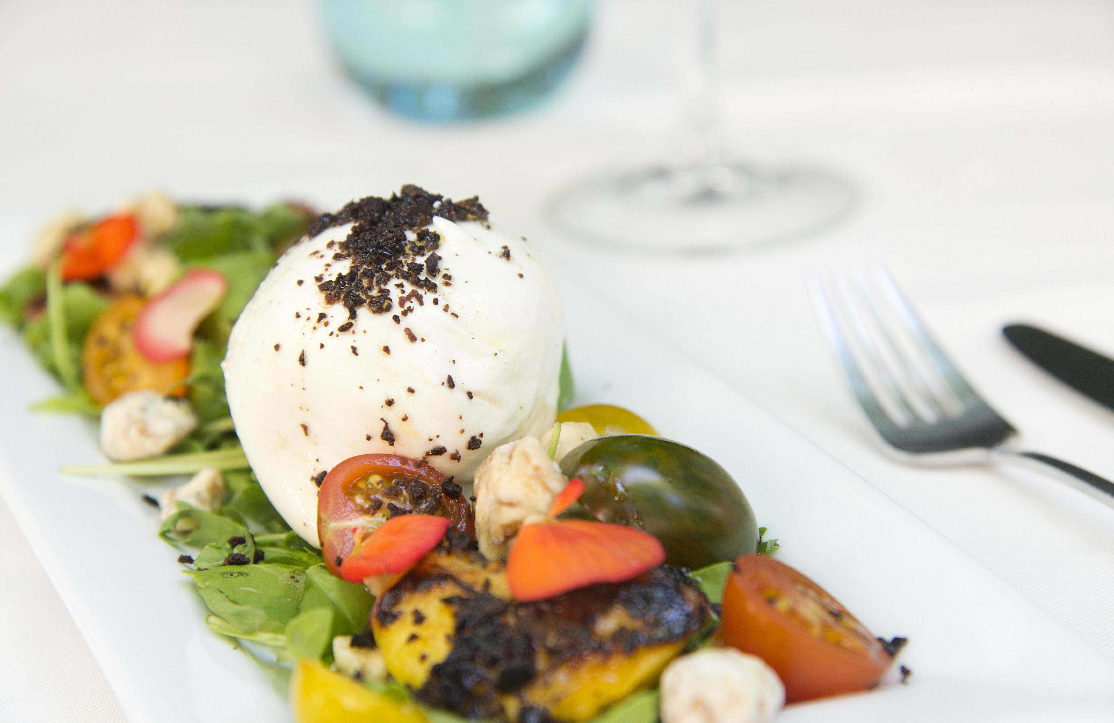 A plated dish with grilled vegetables, a poached egg, and a glass of wine on a white table.
