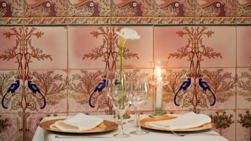 Una mesa servida con platos, vasos y una vela, frente a una pared con un diseño de azulejos florales.