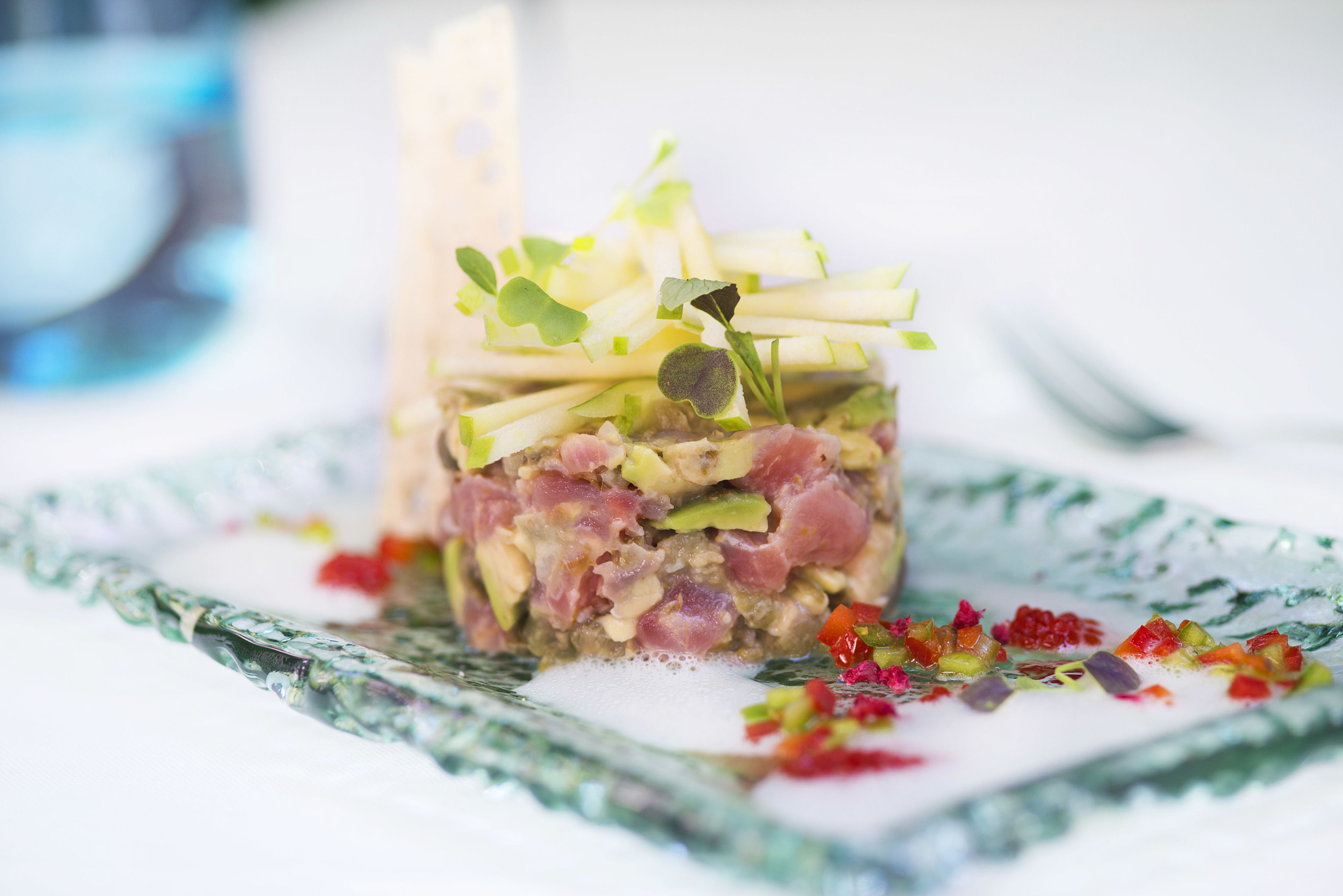 A close-up of a layered dish with various ingredients, garnished with microgreens and edible flowers, on a patterned glass plate.