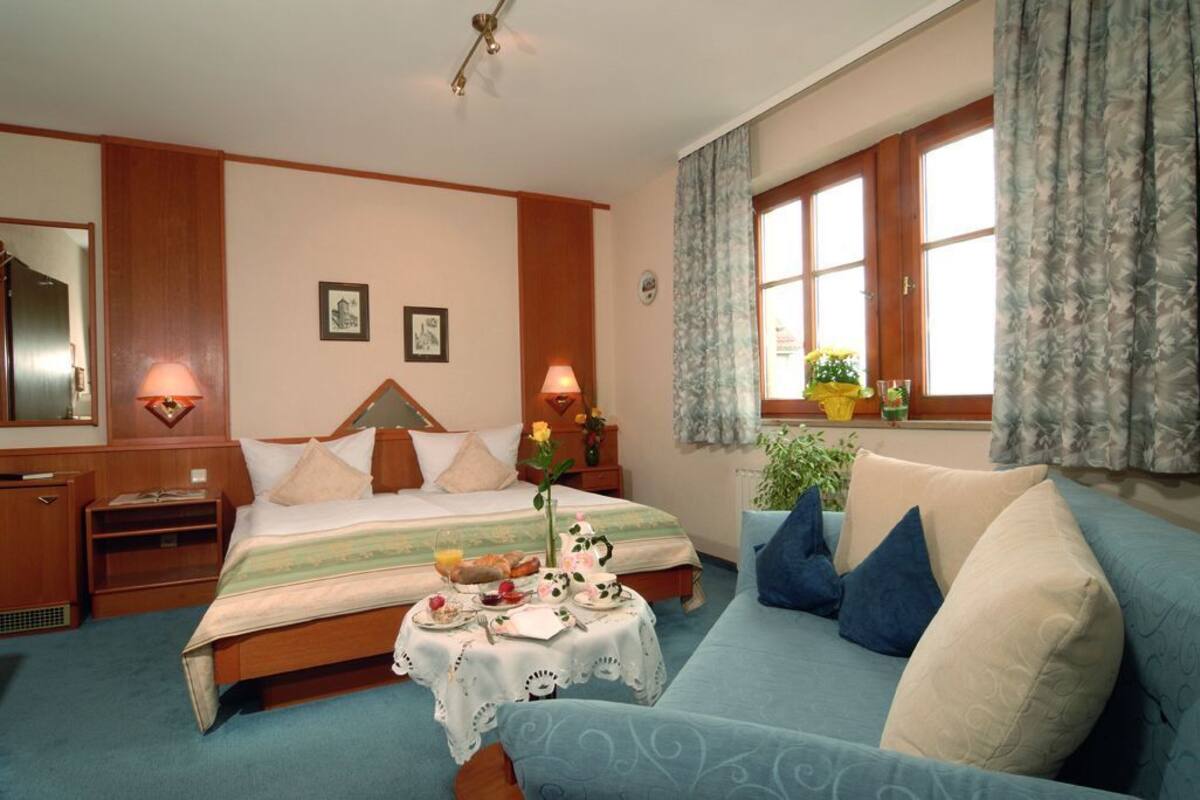 Habitación de hotel con cama, mesitas de noche, ventana con cortinas, sofá y una pequeña mesa con alimentos para el desayuno.