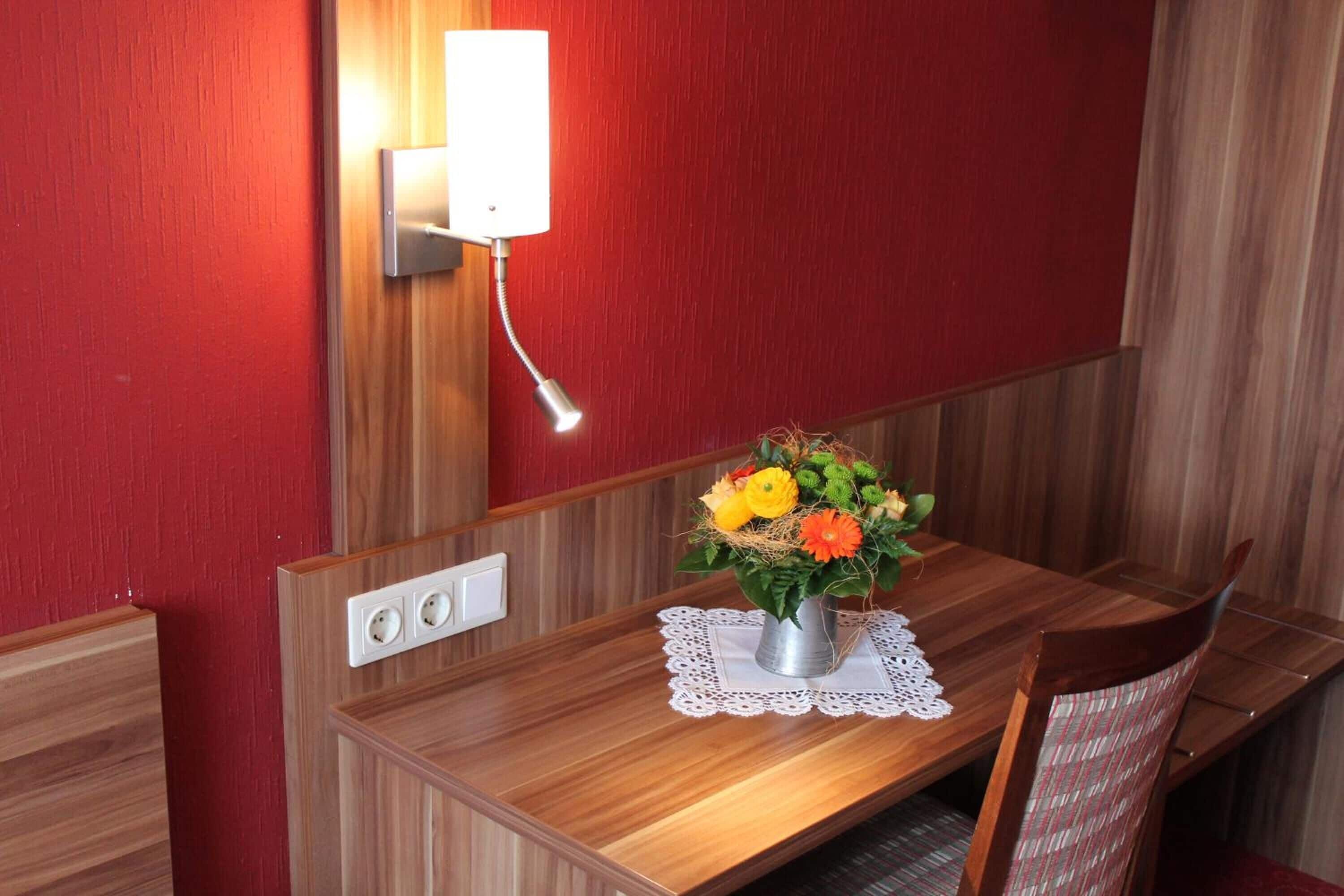 A wooden dining table with a metal vase of flowers, a white doily, and a wall-mounted light fixture.