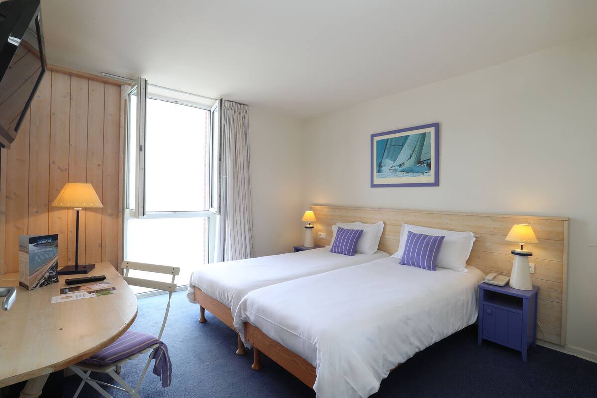 A hotel room with two beds, a wooden headboard, a desk with a lamp, a chair, and a window with curtains.