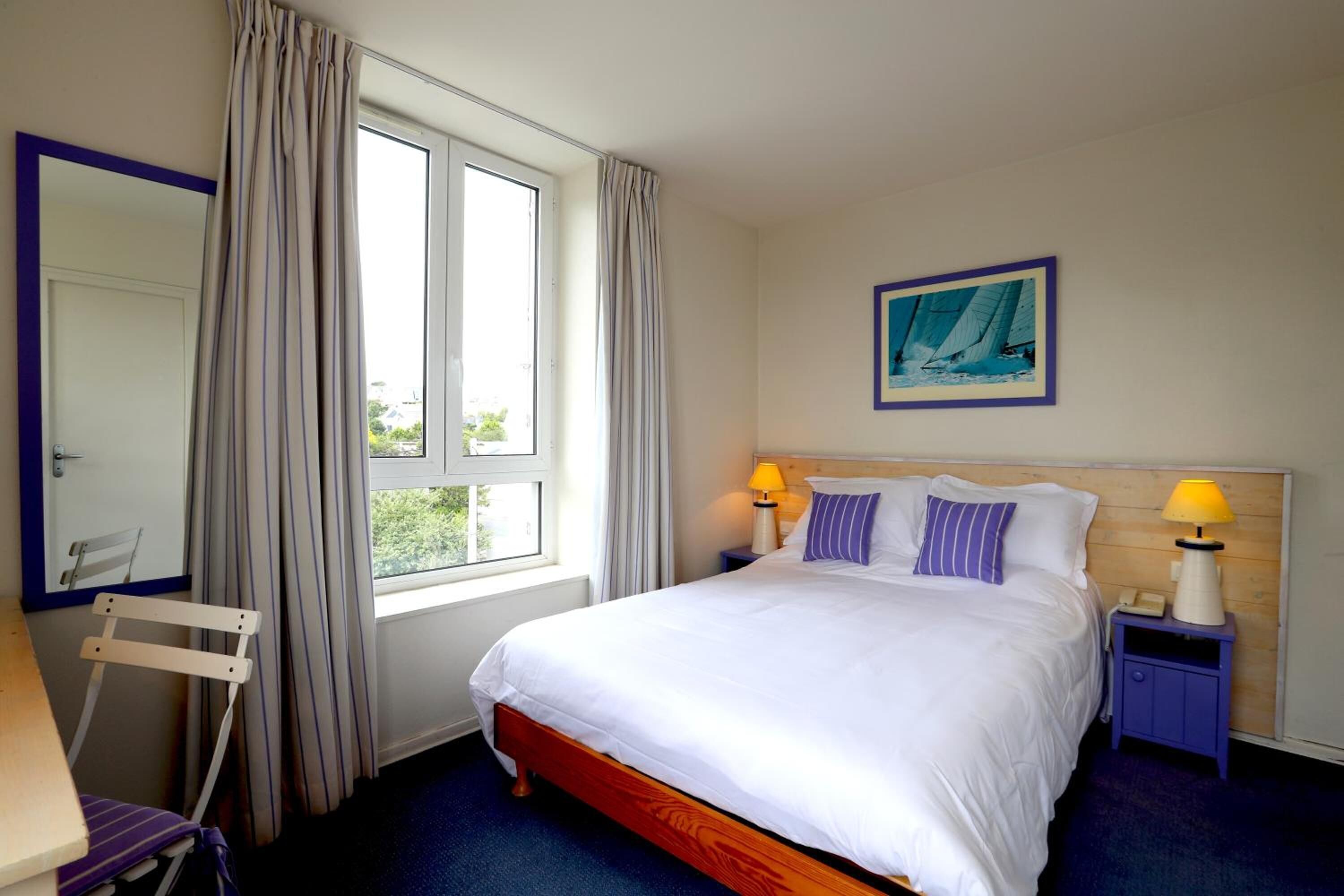 Een hotelkamer met een bed, twee bedlampen, een raam met gordijnen, een stoel, een bureau en een schilderij aan de muur.