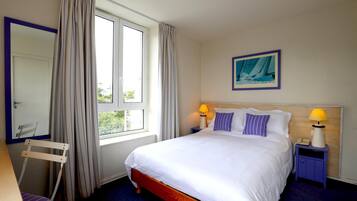 Een hotelkamer met een bed, twee bedlampen, een raam met gordijnen, een stoel, een bureau en een schilderij aan de muur.