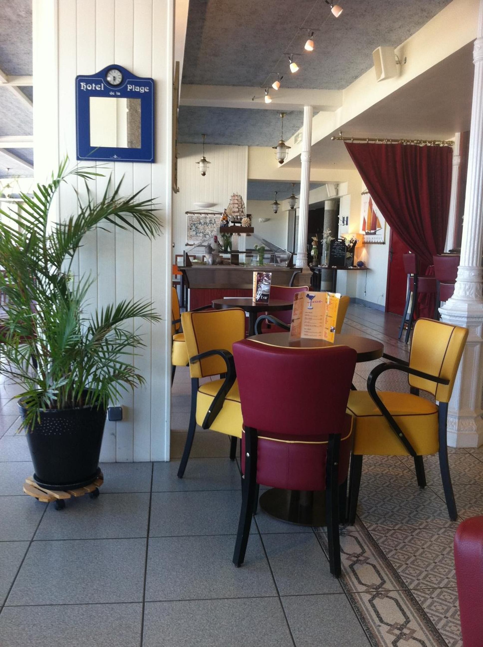 Een hotel lobby met een receptiebalie, gele en rode stoelen, een potplant en een bordje met de tekst 'Hotel de la Plage'.