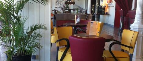 Een hotel lobby met een receptiebalie, gele en rode stoelen, een potplant en een bordje met de tekst 'Hotel de la Plage'.