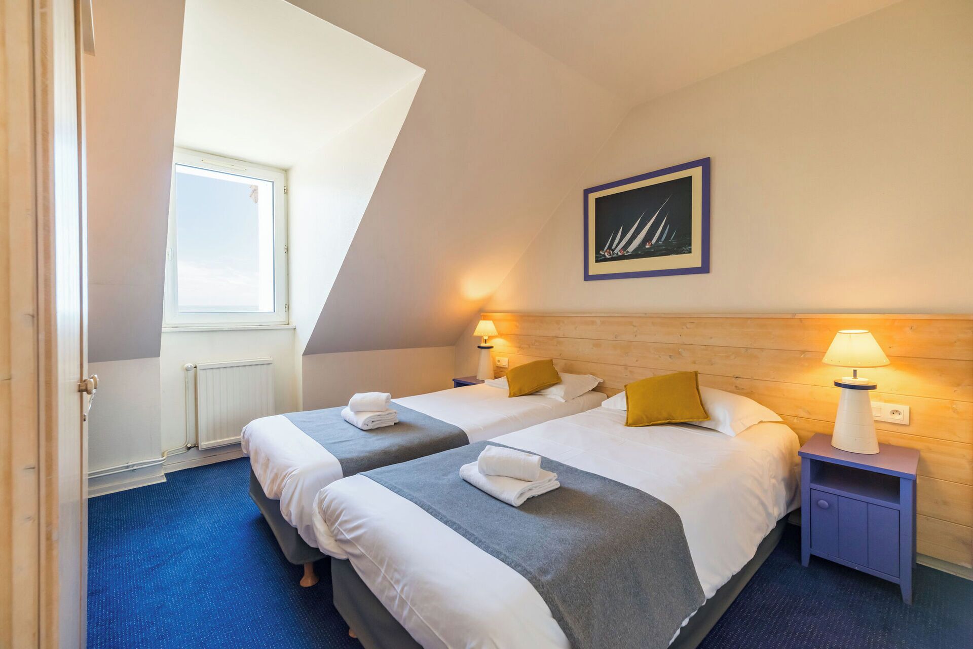 Ein Hotelzimmer mit zwei Betten, einem Fenster, einem gerahmten Bild an der Wand und einem blauen Teppich.