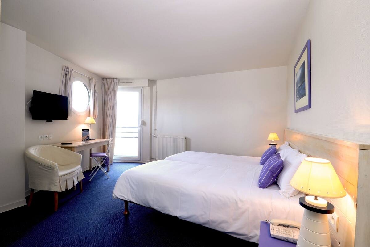 A hotel room with a bed, a desk with a chair, a television, and a window with curtains.