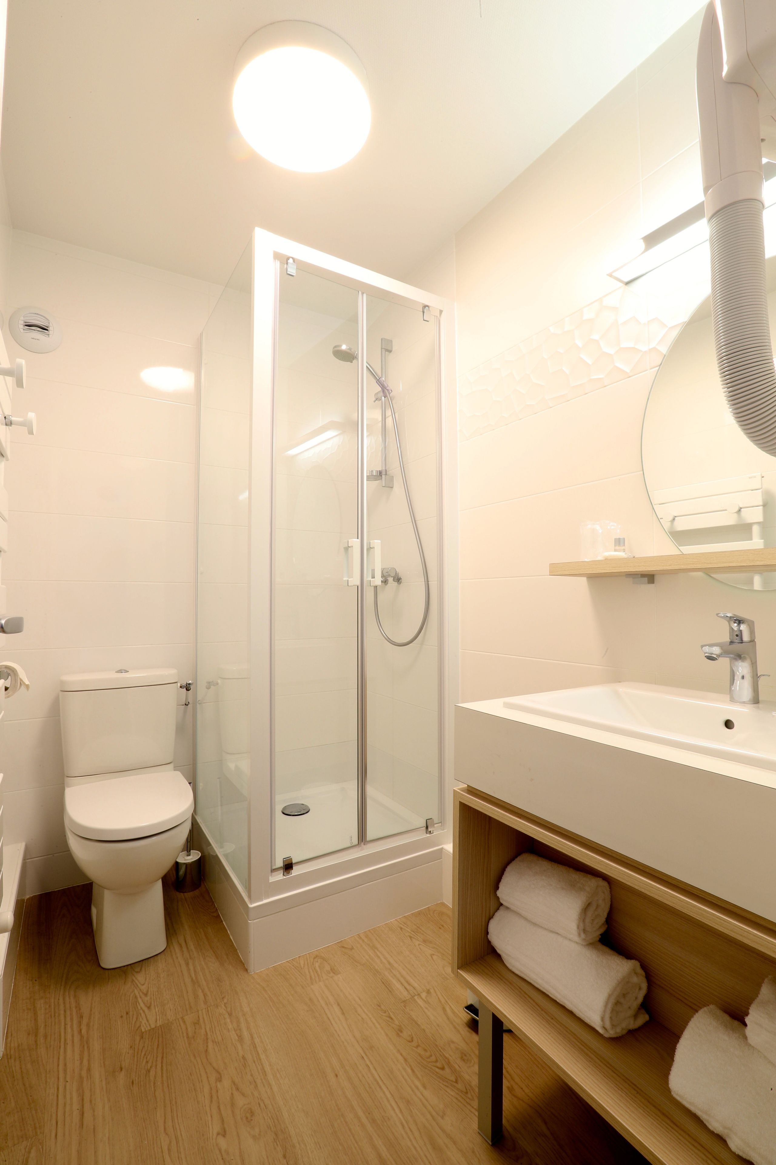 Een moderne badkamer met een douchecabine van glas, een wit toilet, een houten wastafelmeubel met spiegel en een plank met handdoeken.