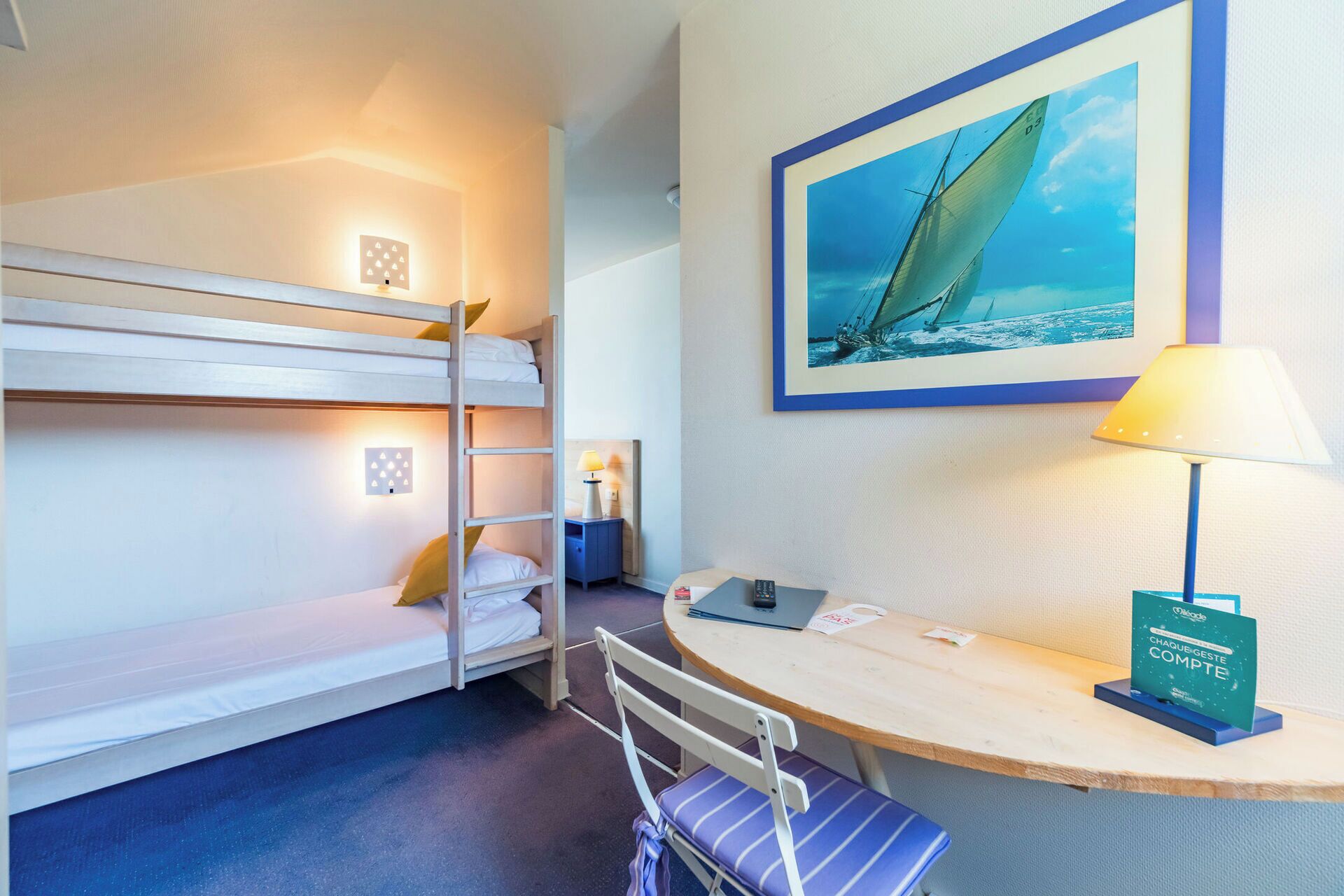Et hotelværelse med køjesenge, et skrivebord med stol, en lampe, et indrammet billede af en sejlbåd og en skilt til concierge.