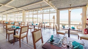 A restaurant with tables set for dining, large windows offering a view of the beach, and a ceiling with exposed beams. - Hôtel Miléade de la Plage - Binic (Binic-Etables-sur-Mer)