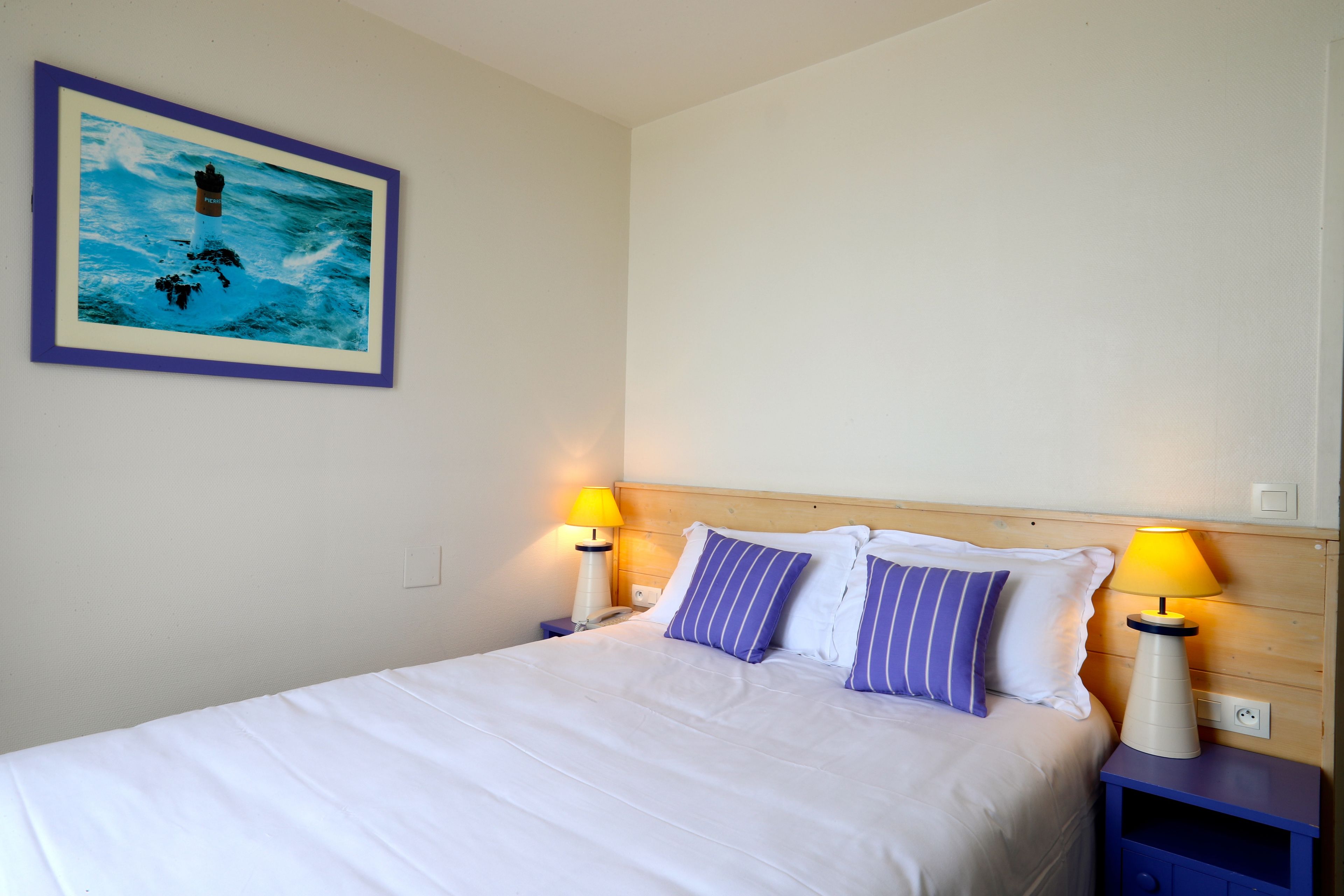Una cama bien hecha con sábanas blancas, dos mesitas de noche con lámparas y un cuadro con un faro en la pared.