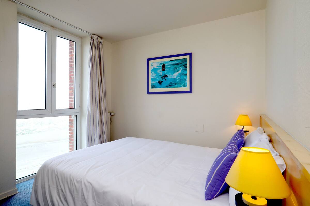 A hotel room with a bed, a window with curtains, a wall-mounted picture, and a bedside lamp.