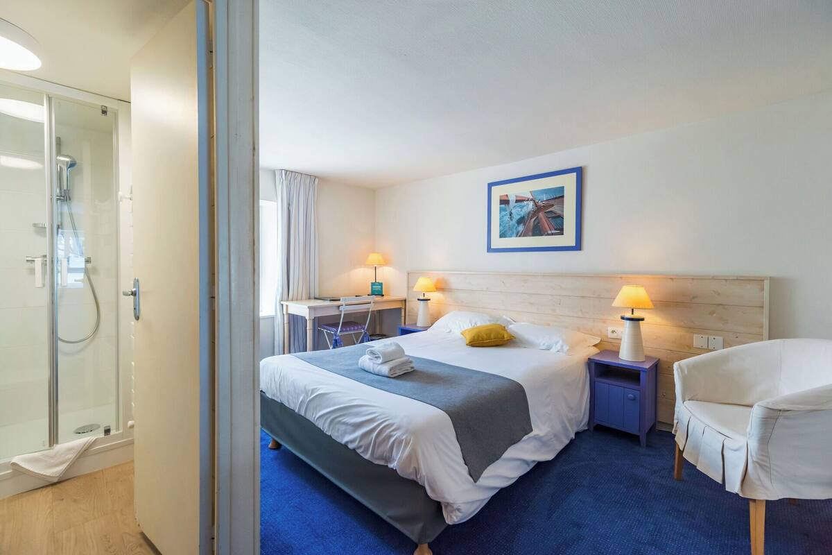 A hotel room with a bed, two bedside tables with lamps, a chair, a shower area, and a painting on the wall.