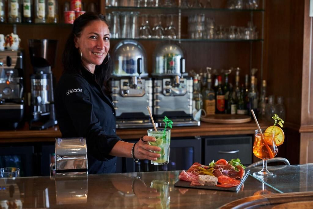Eine Bar mit einer Person, die ein Getränk hält, einem Glas mit Zitronenzeste und einer Speisenauswahl auf einem Tablett.
