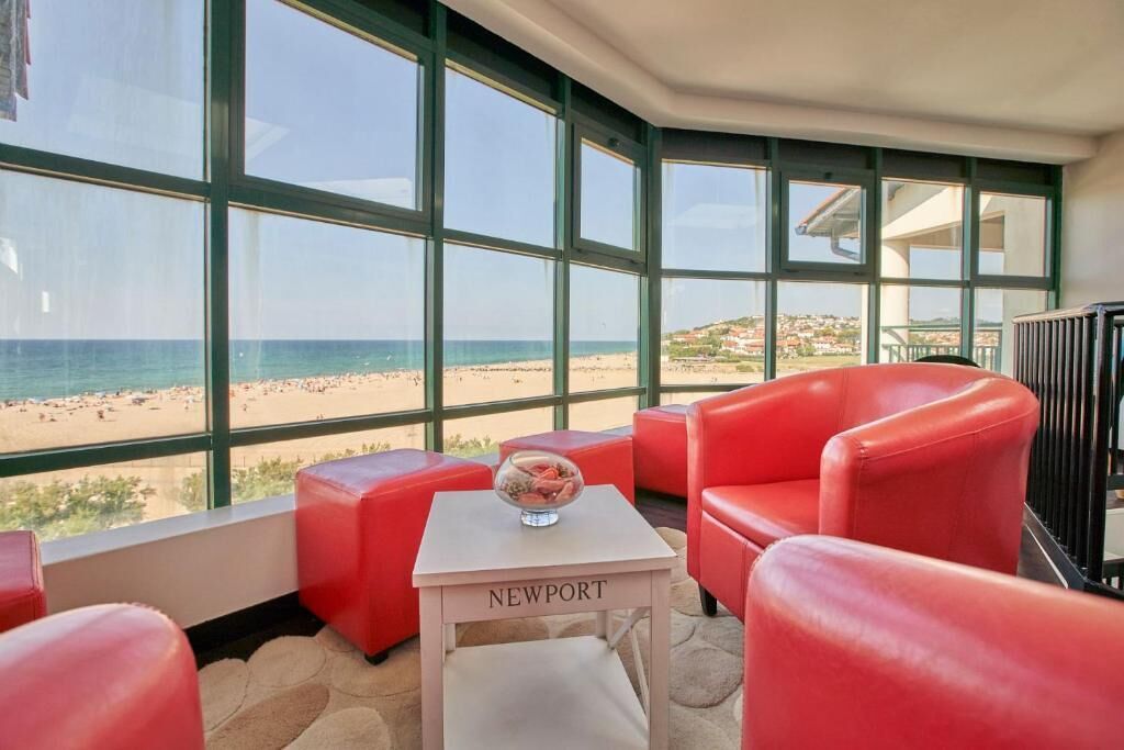 A room with a large window overlooking a beach, red leather chairs, a small white table with a bowl, and a sign that reads 'NEWPORT'.