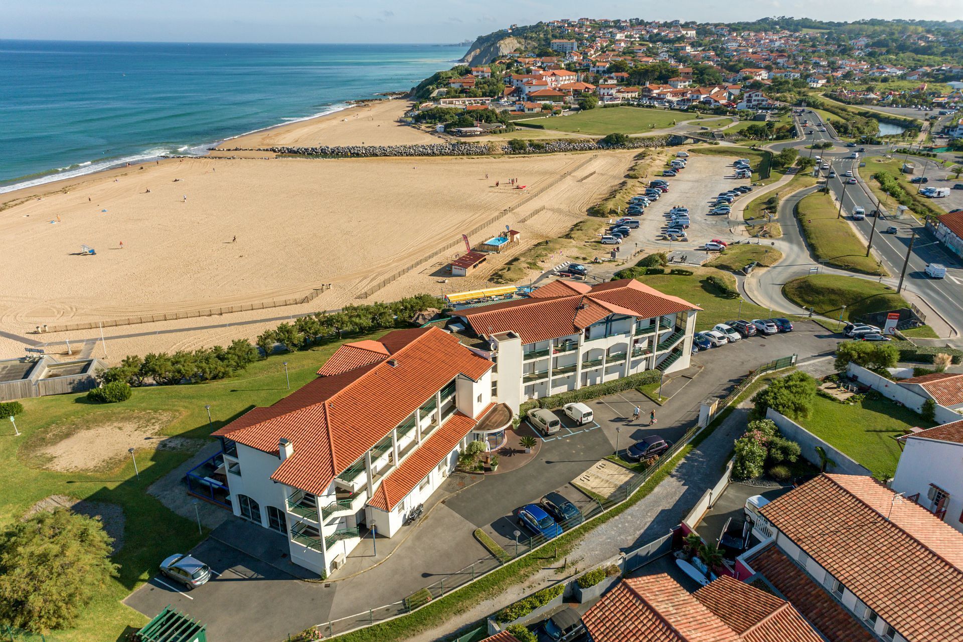 Vista aérea de una zona costera con una playa, un estacionamiento y un edificio de varios pisos con techo de tejas rojas.