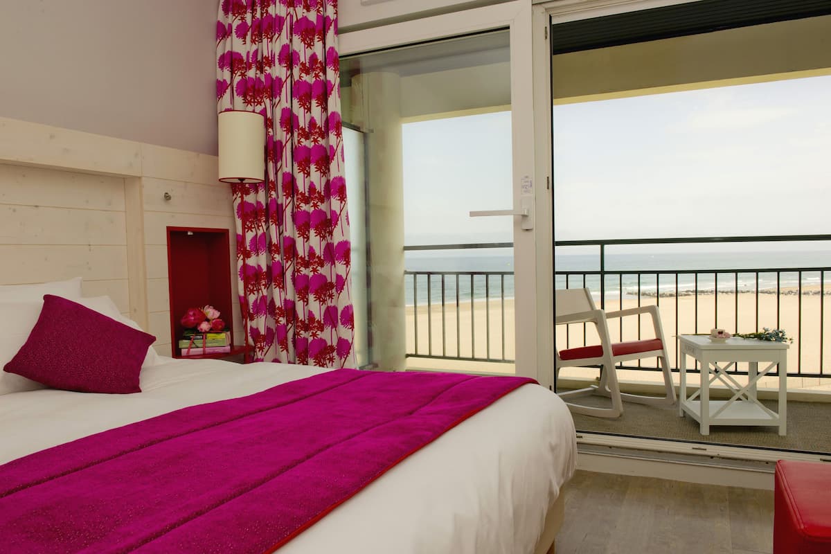 A hotel room with a bed, a balcony with a view of the beach, a chair, and a small table.