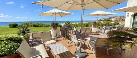 Eine Terrasse im Freien mit mehreren Tischen und Stühlen, großen Sonnenschirmen und Blick aufs Meer.