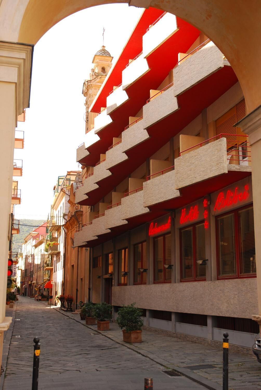 A narrow street with a building featuring a red and white facade, a visible restaurant sign, and a bell tower in the background.