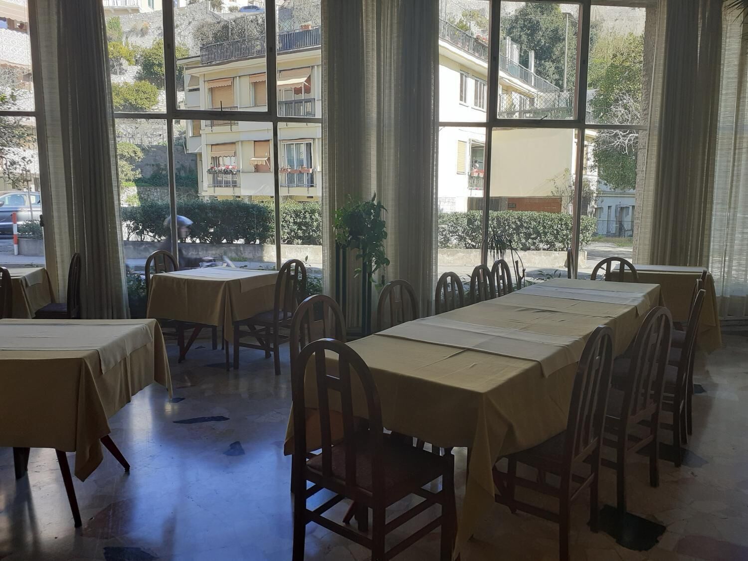 A dining area with tables and chairs, large windows with curtains, and a view of buildings and greenery outside.
