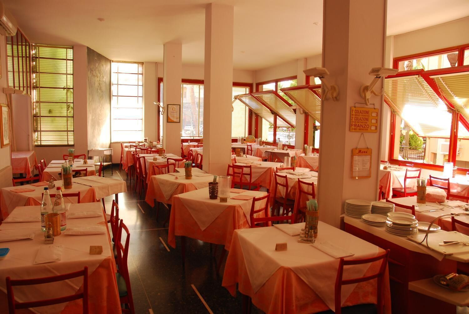 A restaurant with multiple tables and chairs, large windows with blinds, and a menu board on the wall.