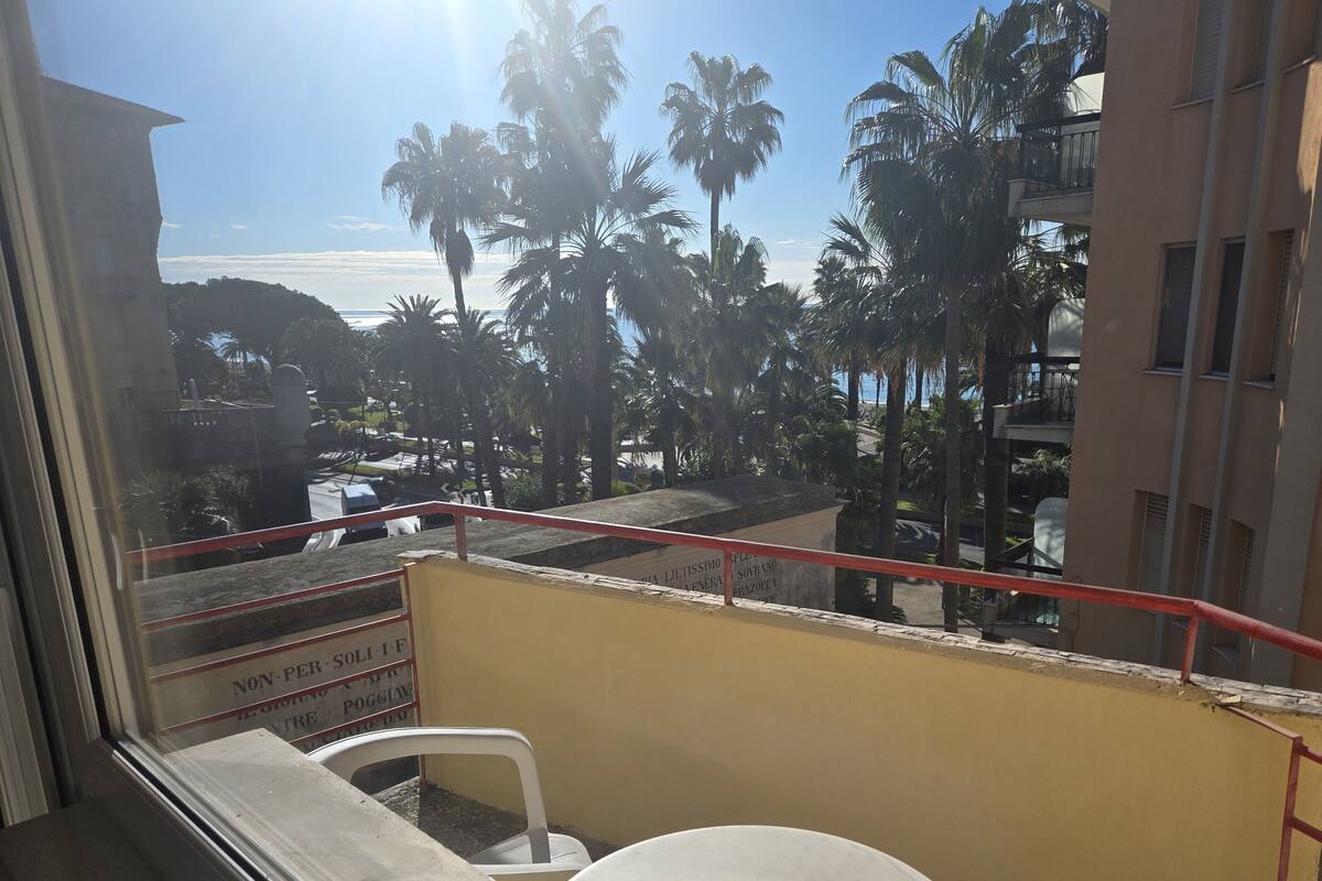 A balcony with a view of palm trees and a beach, a white chair, and a small table.