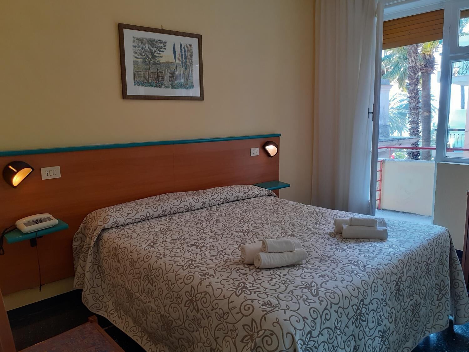 A hotel room with a bed, a bedside table, a phone, a framed picture on the wall, and a window with a view of palm trees.