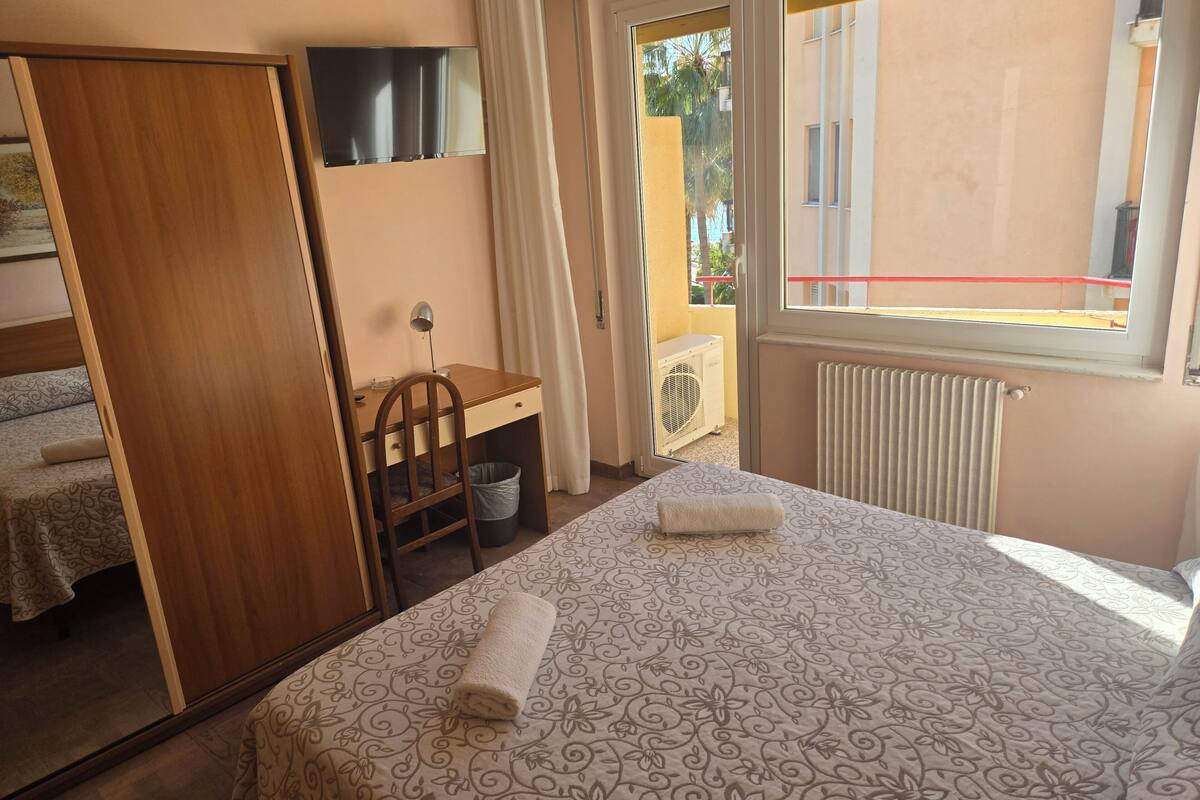 A bedroom with a bed, a desk, a chair, a radiator, and a window with a view of the outside.
